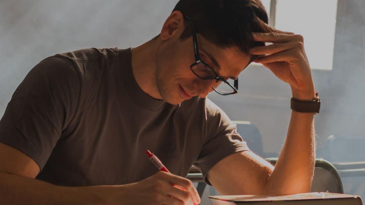 A young man with glasses intensely focused on writing in a notebook in a dim classroom.