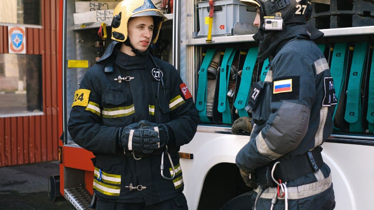 Two firefighters in full protective gear converse by a fire truck.