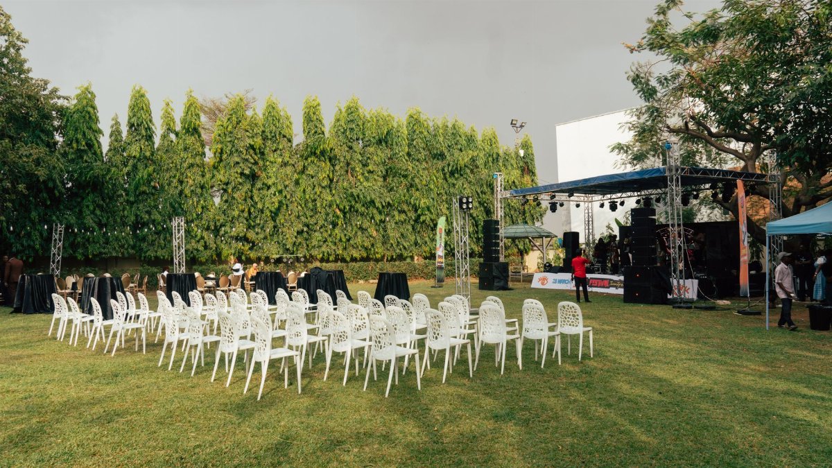 An outdoor event venue with white chairs, stage setup, sound equipment, and greenery surrounding the space.