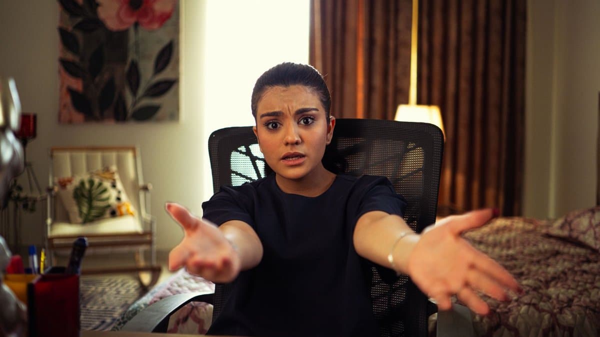 A young woman seated indoors, extending arms in an expression of frustration.