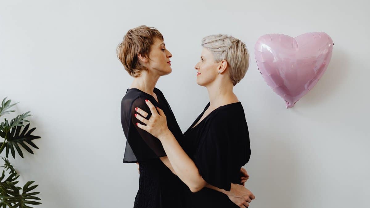A loving lesbian couple embraces indoors with a pink heart-shaped balloon symbolizing lgbt pride.