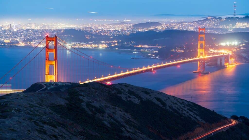 Capture of the illuminated Golden Gate Bridge at dusk with San Francisco skyline in the background.