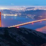 Capture of the illuminated Golden Gate Bridge at dusk with San Francisco skyline in the background.