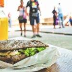 Close-up of a sandwich and orange juice on a table with people in the park.