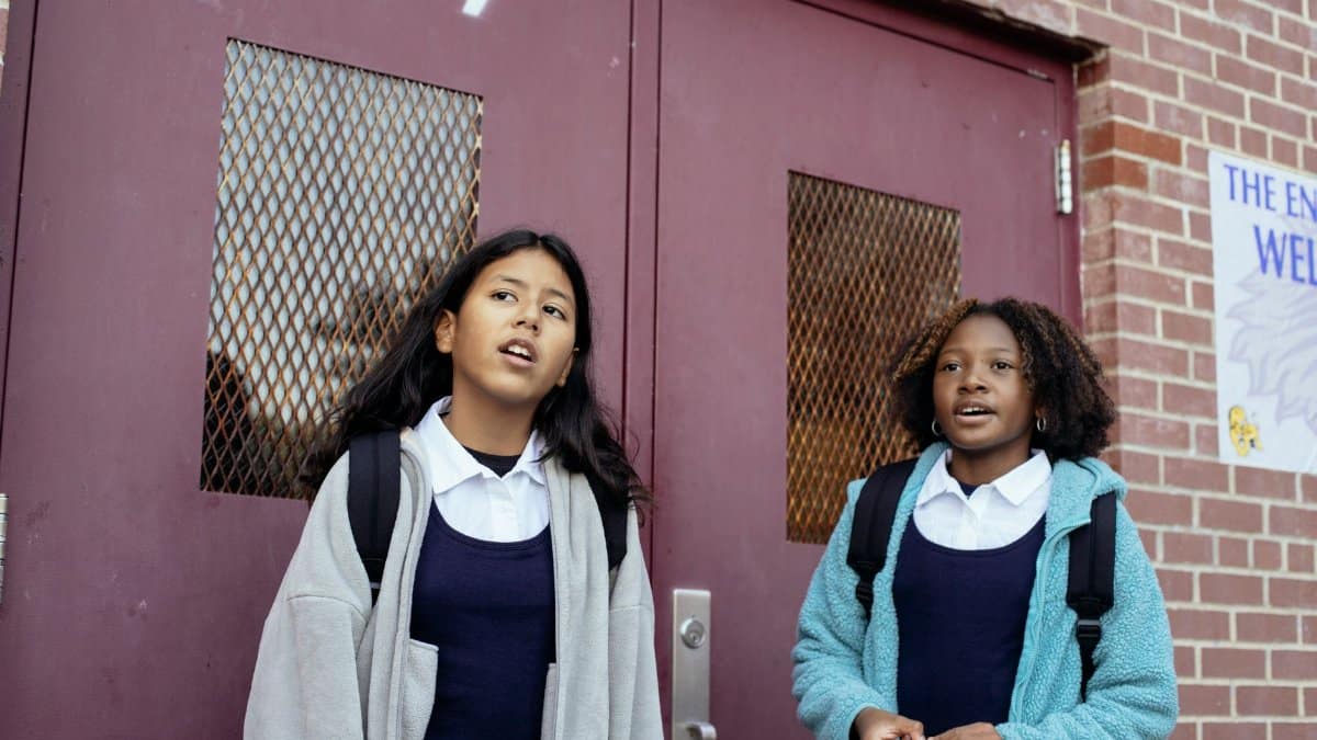 Positive multiracial girls classmates in warm jackets standing together near masonry school wall and looking away