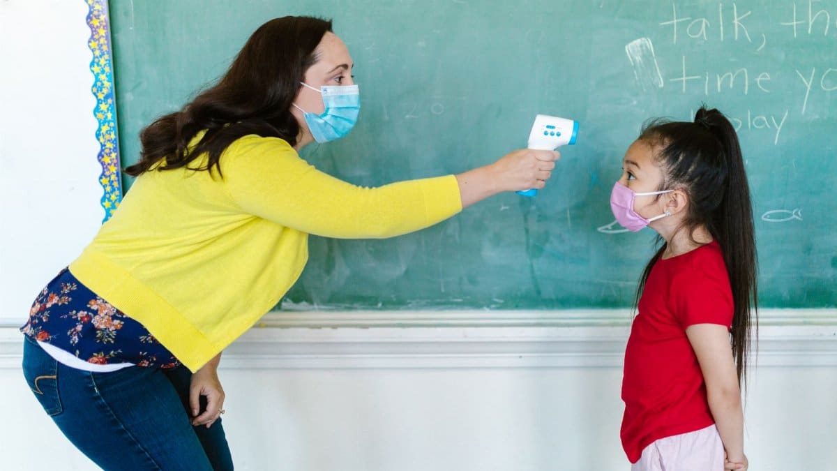 A teacher checks a student's temperature in a classroom setting, emphasizing school safety protocols with face masks.
