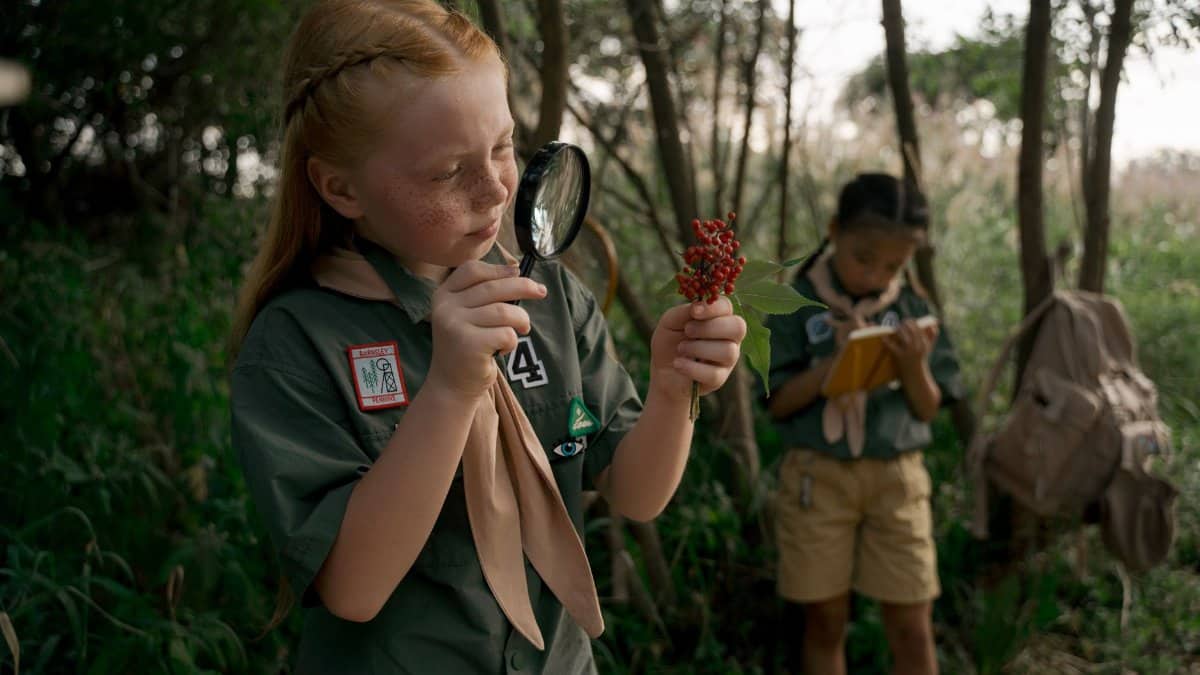 Children exploring nature at a summer scouting camp, using magnifying glasses and taking notes.