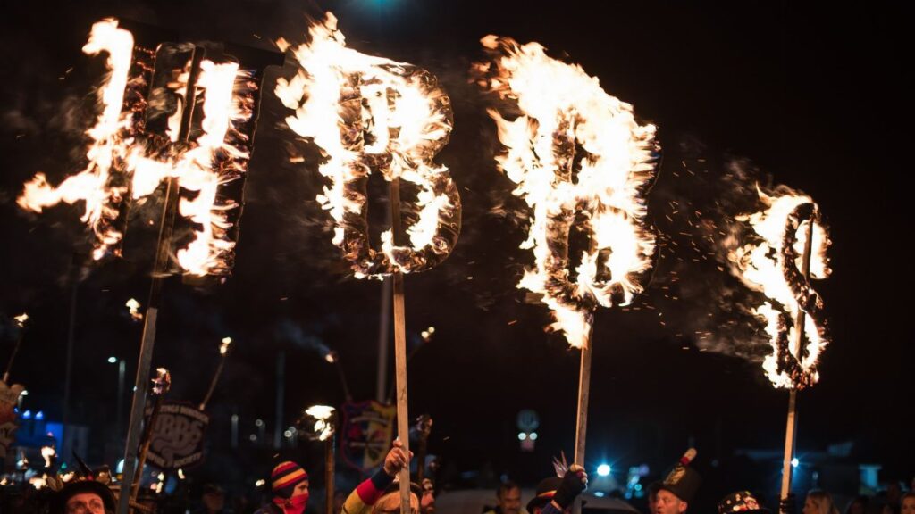 Dramatic torchlight parade at night featuring burning letters and colorful attire in a festive atmosphere.