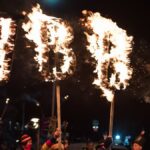 Dramatic torchlight parade at night featuring burning letters and colorful attire in a festive atmosphere.