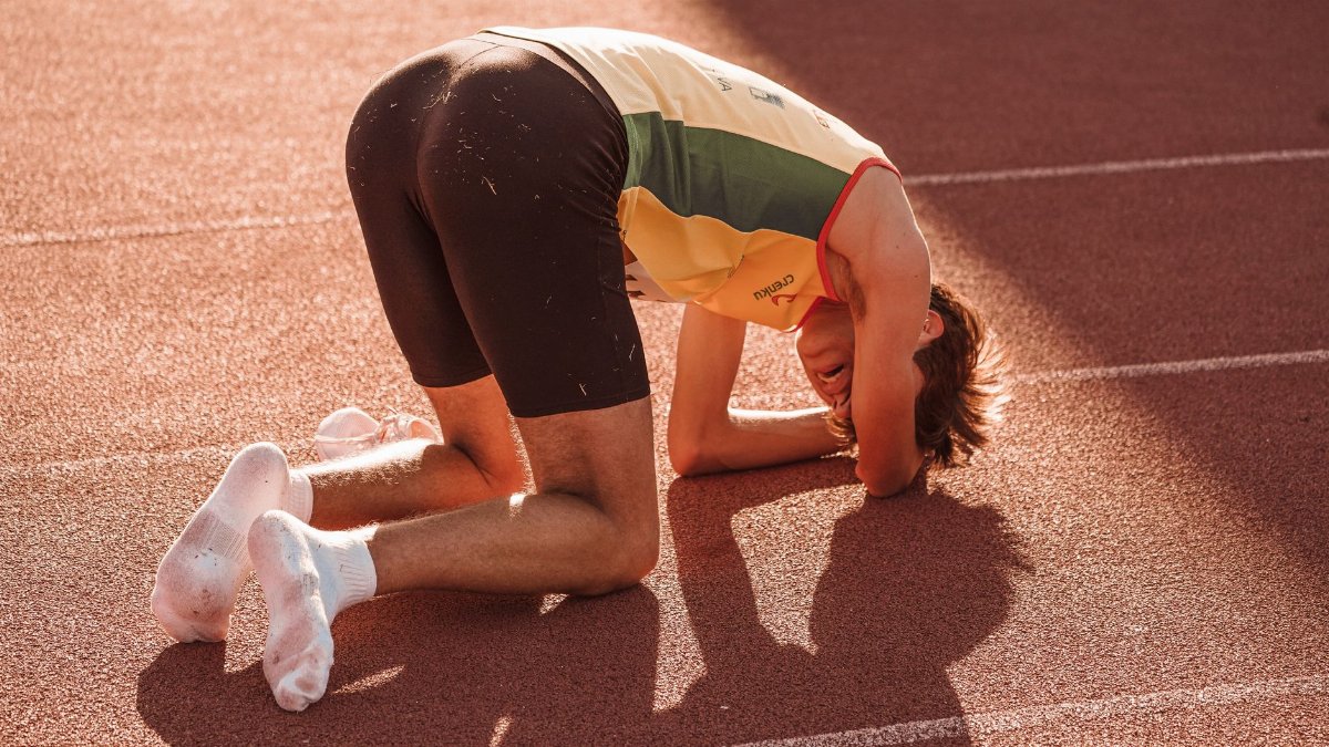 An athlete in sportswear kneels exhausted on a sunlit track, reflecting fatigue and determination.