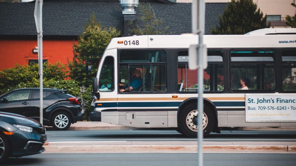A city bus passing through an urban street with cars and buildings in the background.