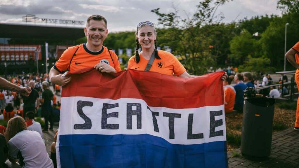 Two soccer fans in orange jerseys holding a Seattle flag outside a stadium in Dortmund.