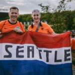 Two soccer fans in orange jerseys holding a Seattle flag outside a stadium in Dortmund.