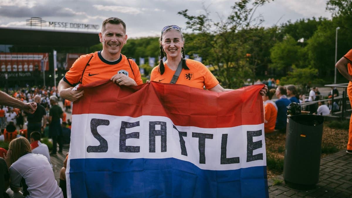 Two soccer fans in orange jerseys holding a Seattle flag outside a stadium in Dortmund.