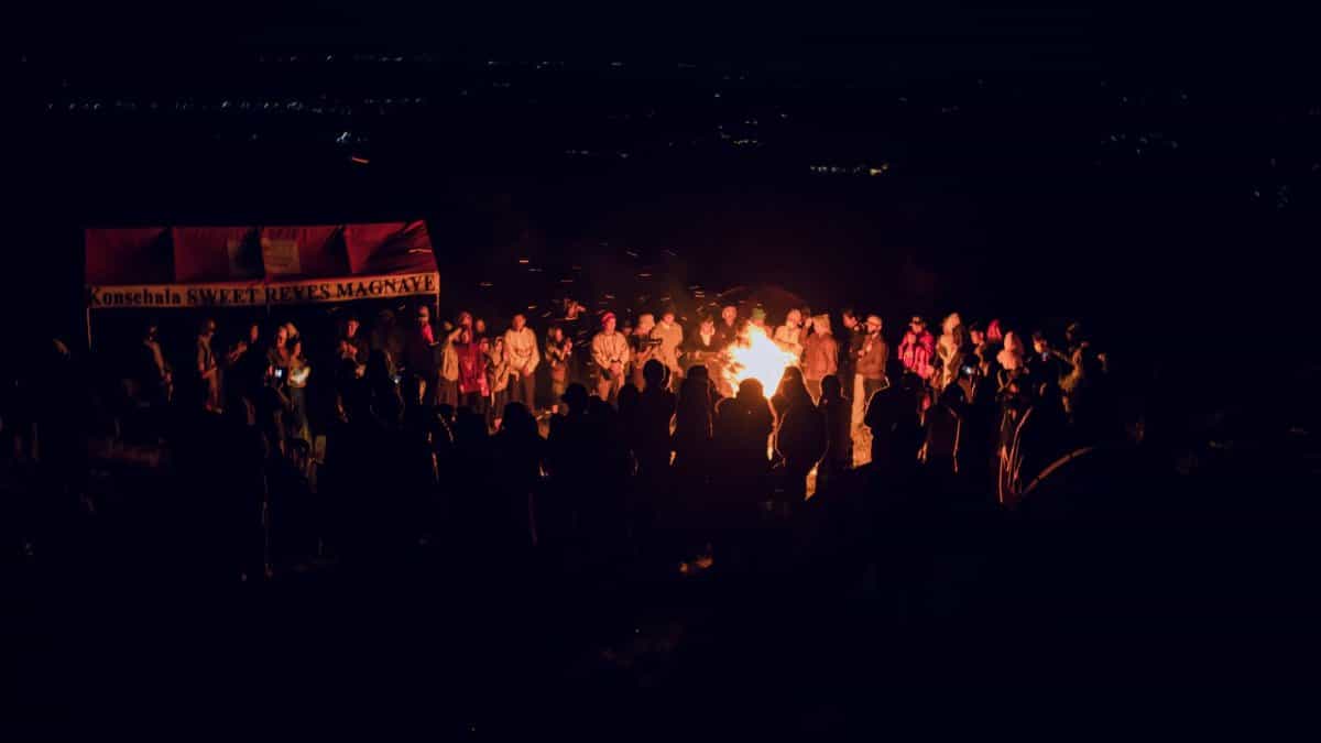 A lively group gathers around a bonfire during an outdoor night festival, celebrating under the stars.