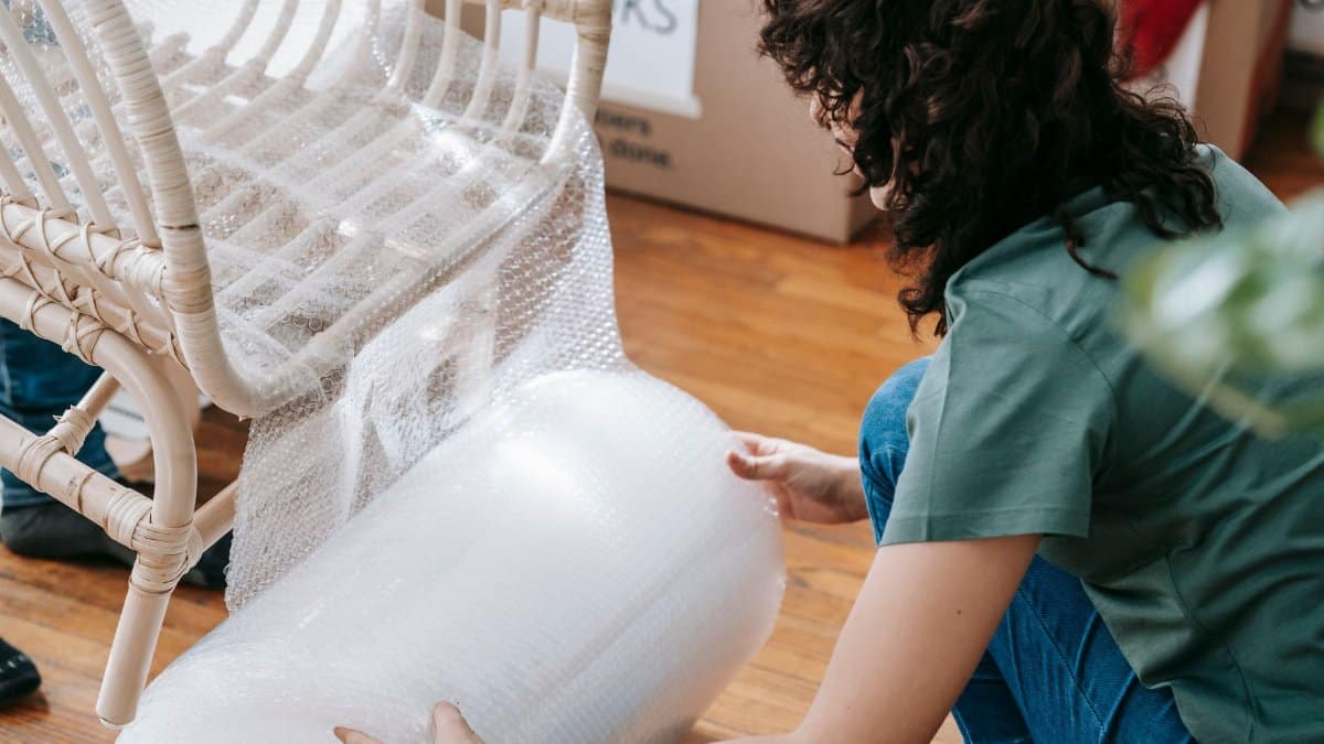 Woman kneeling to wrap a wicker chair with bubble wrap, preparing for relocation.