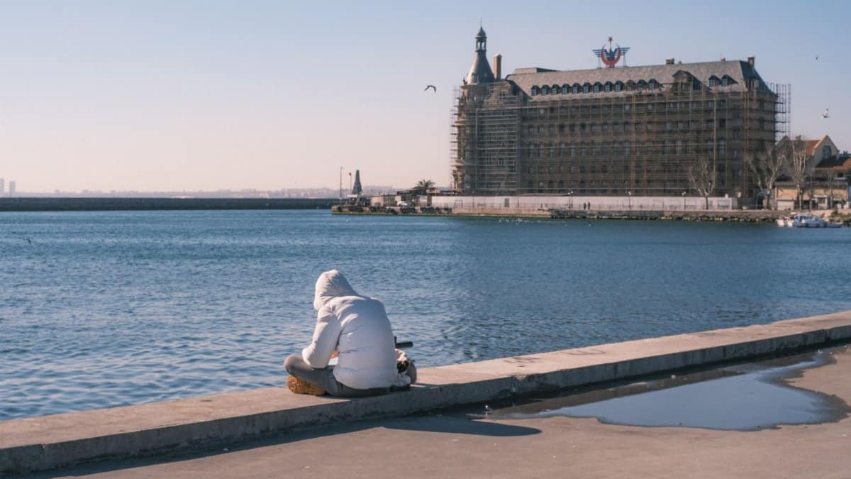 A person in a white jacket sits by a waterfront facing a historic building.