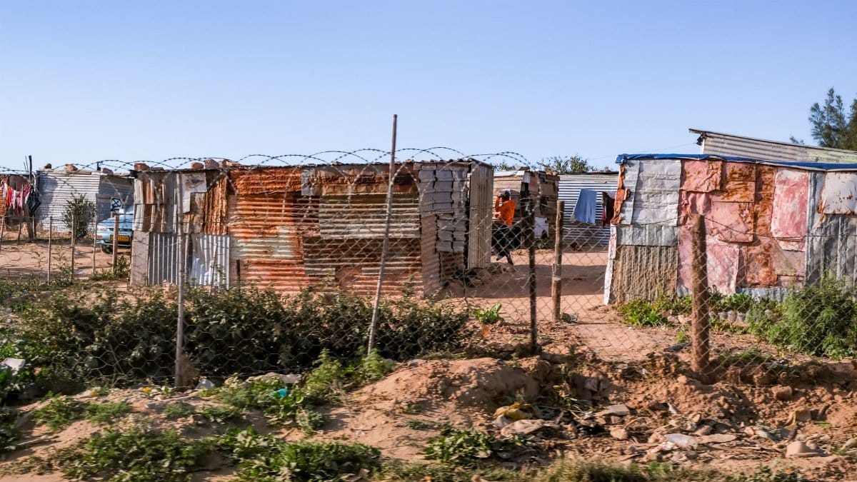 Outdoor view of a rural shantytown with metal shacks and barbed wire fencing under a clear sky.
