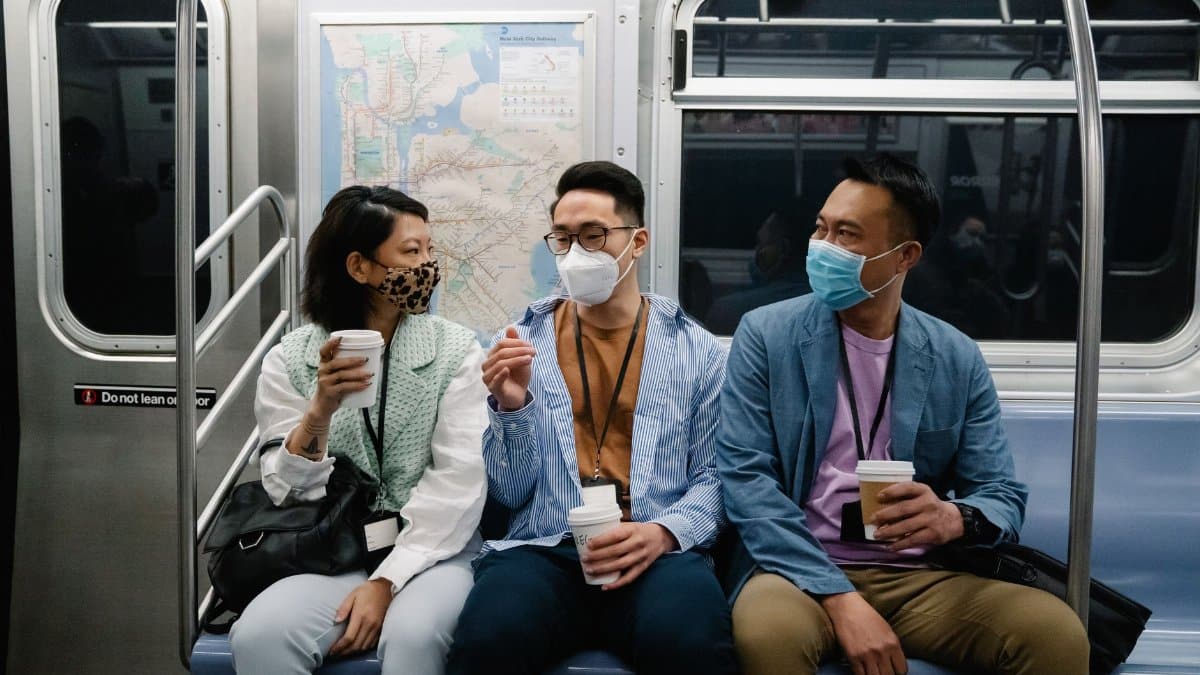 Three masked commuters sitting together on a subway train, holding coffee cups.
