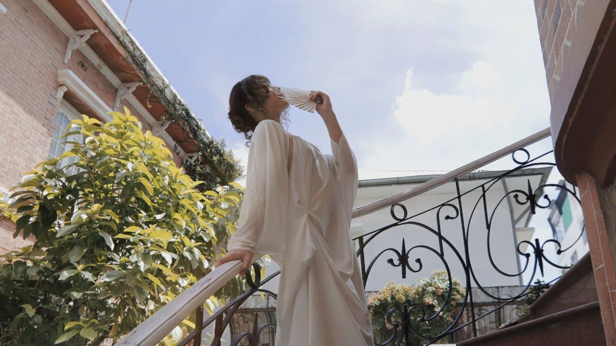 A woman in a flowing dress gracefully poses with a fan on an outdoor staircase under clear blue skies.