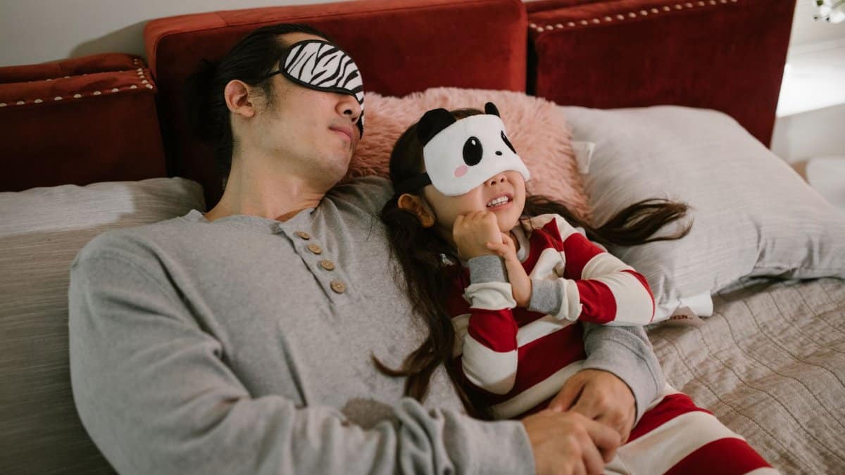 A father and daughter wearing sleep masks relax on a cozy bed, enjoying a moment together.