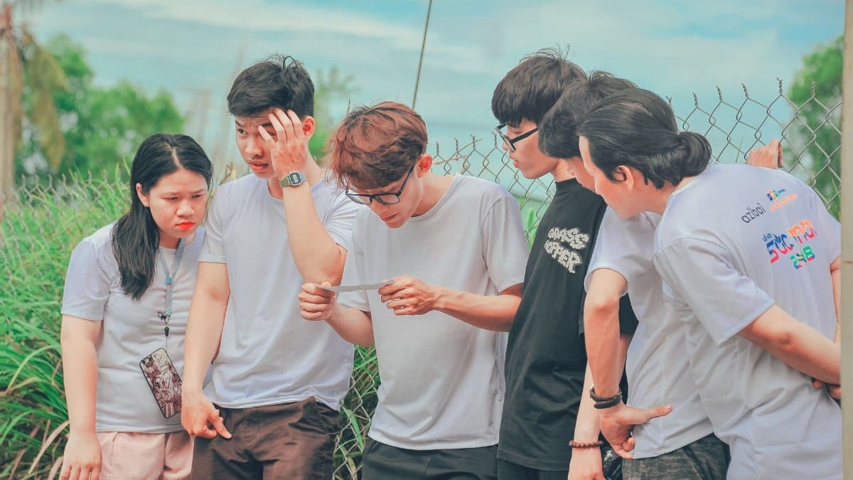 A group of young adults examining a photo together outdoors by a fence on a sunny day.