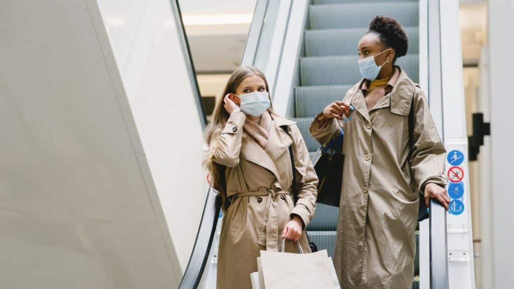 Two women in coats wearing face masks with shopping bags on an escalator indoors.