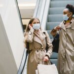 Two women in coats wearing face masks with shopping bags on an escalator indoors.
