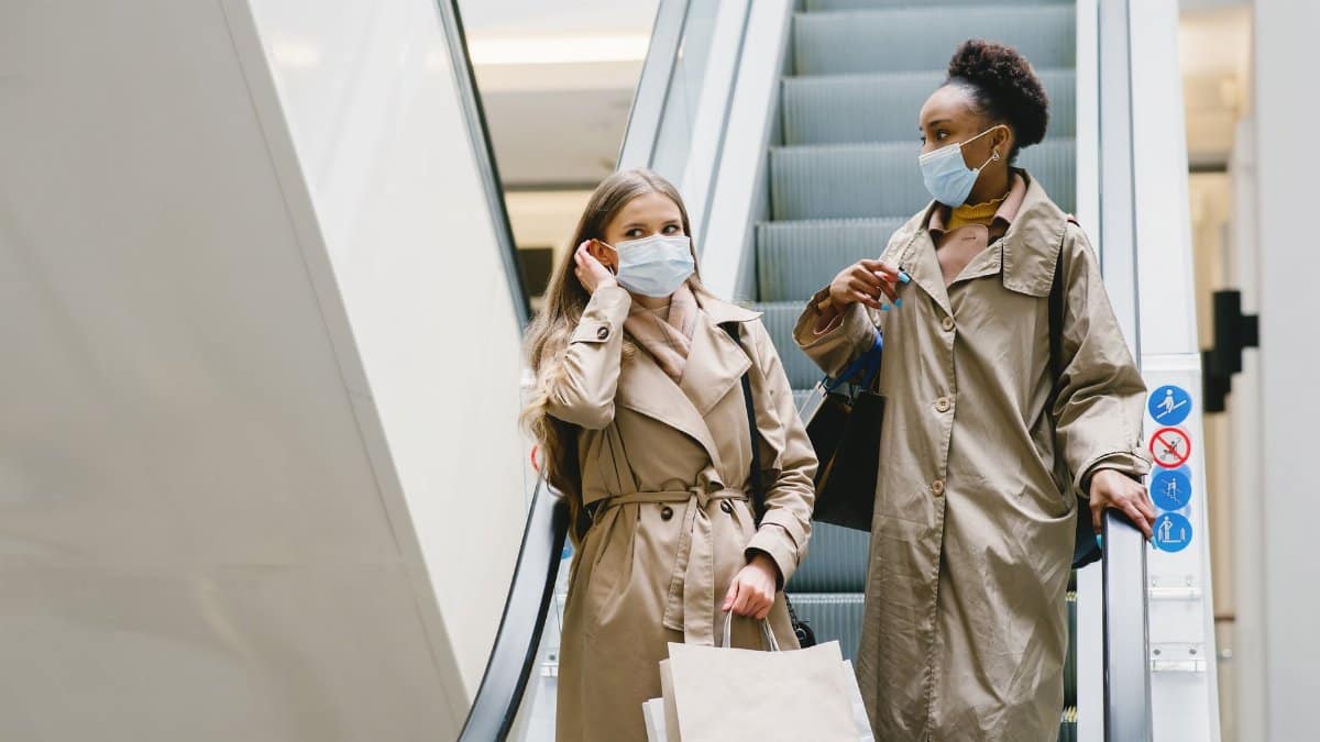 Two women in coats wearing face masks with shopping bags on an escalator indoors.