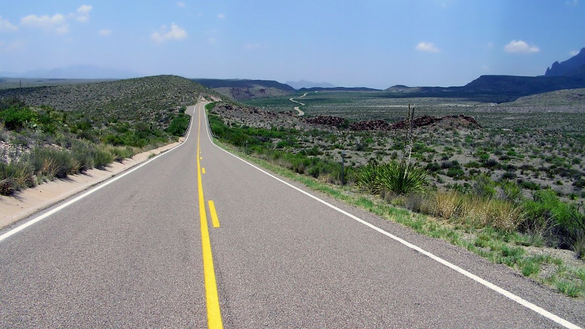 A long open road stretches through a scenic desert landscape under a bright blue sky.