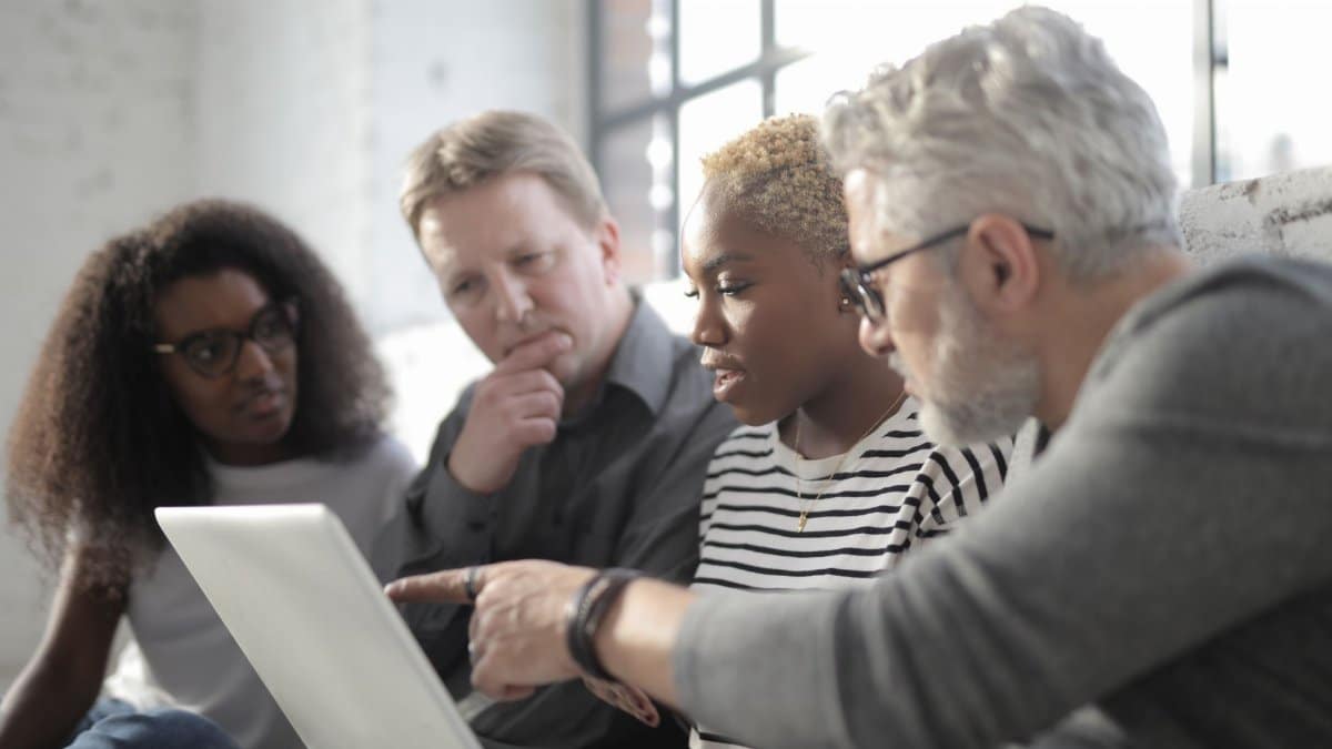 Middle aged employee in eyeglasses pointing at netbook screen with finger while sitting near multiracial pensive colleagues near window in afternoon