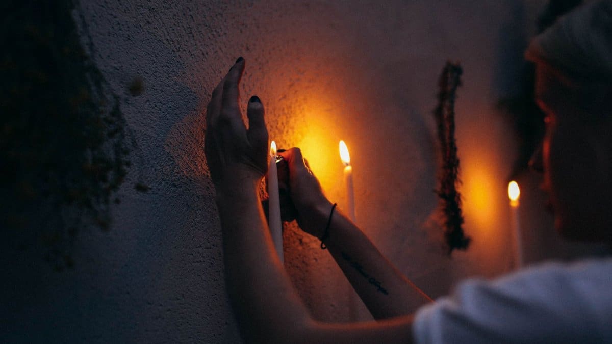 Crop female with lighter burning candlesticks placed near white wall decorated with wreaths in dark room