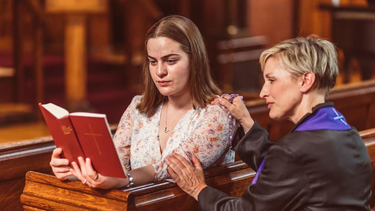 Two women in church, one holding a Bible, engage in thoughtful spiritual reading and prayer.