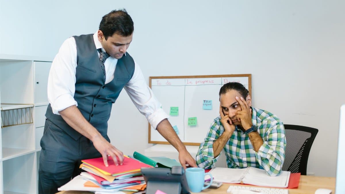 Two businessmen in an office, one stressed at the desk while another points at folders, discussing workload.