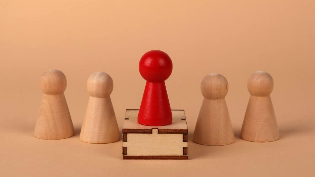 A red pawn stands out as a leader among wooden pieces on a peach background.