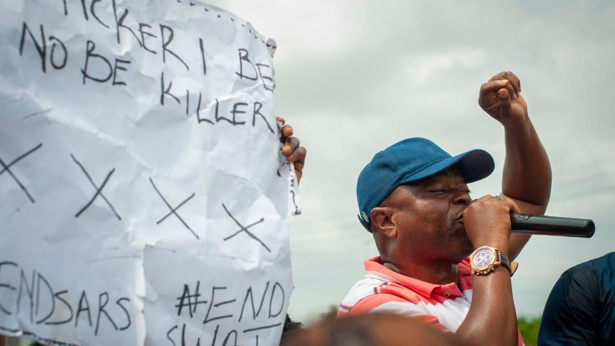 A man passionately speaks through a microphone during a protest, holding a sign with slogans.