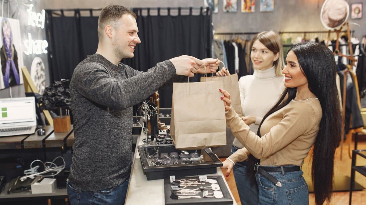 Side view of cheerful male shop assistant standing at counter desk and giving to positive female customers paper bags with purchases in trendy shop