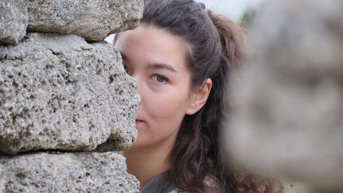 A woman with long hair peers past a stone wall, partially hiding her face, in an outdoor setting.