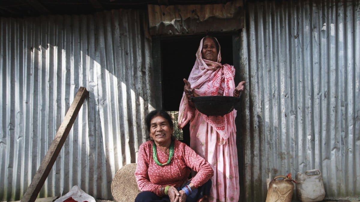 Two elderly women smiling and standing outside a rustic metal house, engaged in daily activities.