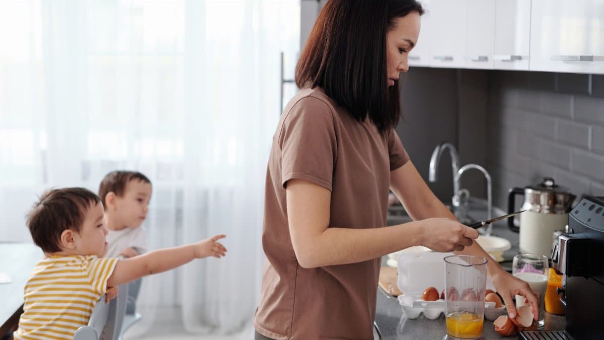 A mother cooks breakfast in a modern kitchen while her twin sons play nearby.