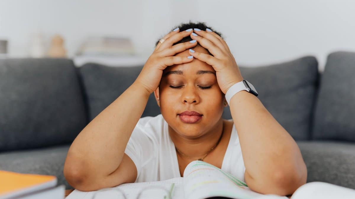A woman with eyes closed holds her head in stress, surrounded by books in an indoor setting.