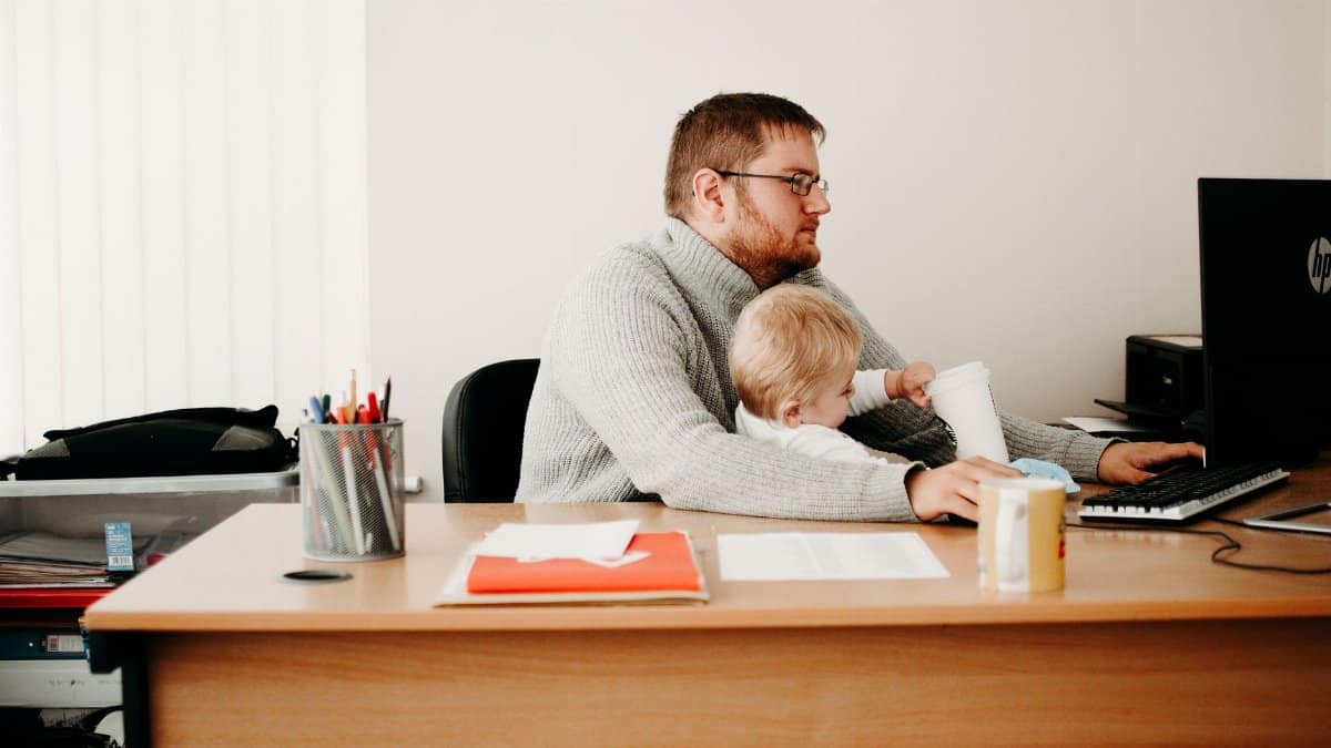 A father multitasks at his desk while holding his child, illustrating remote work and parenting.