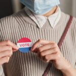 A person wearing a mask holds a voting badge, symbolizing participation in the election.