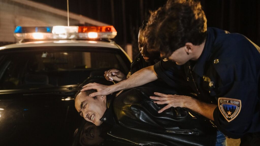Dramatic nighttime arrest scene with police officers detaining a suspect on a patrol car.