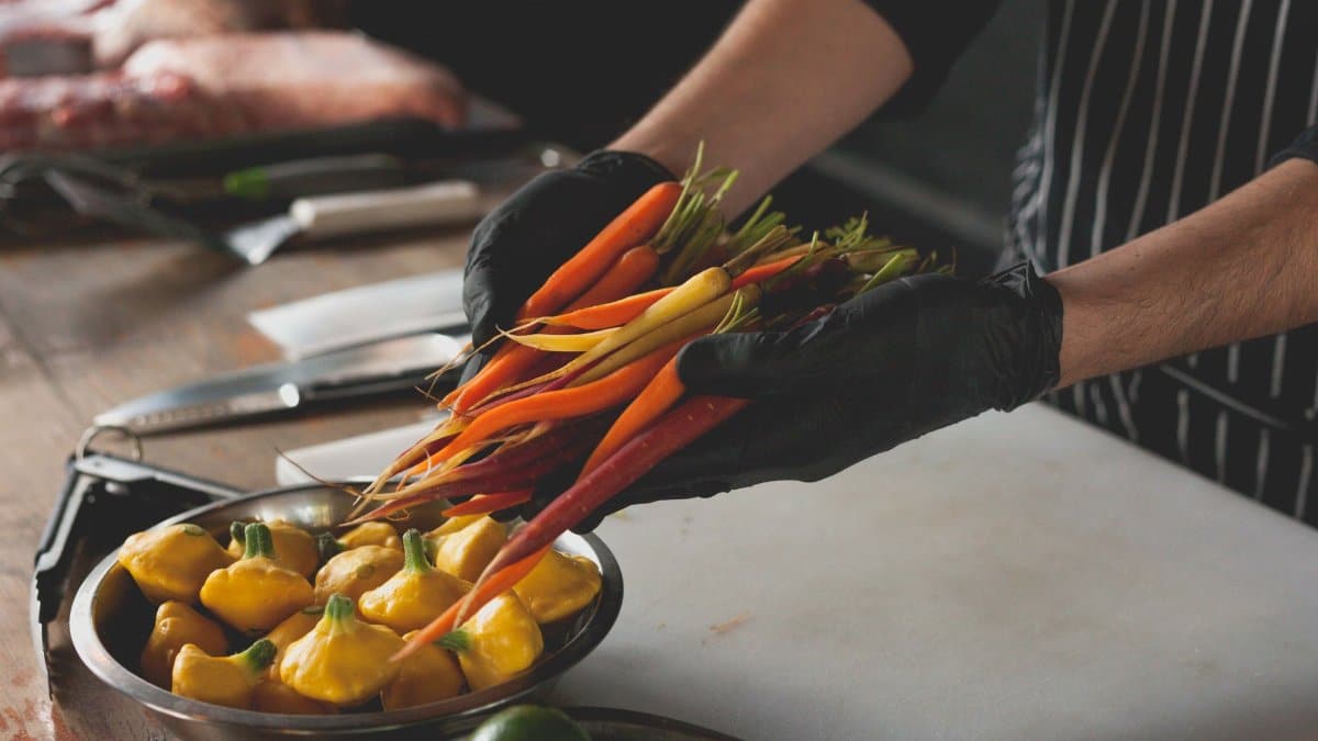 Chef wearing gloves handles fresh carrots and patty pan squash, highlighting preparation