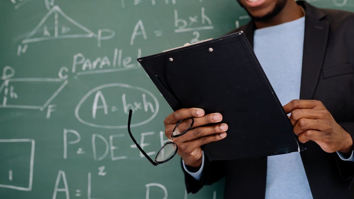 Smiling teacher holding a clipboard in front of a math-filled chalkboard, creating an educational atmosphere.