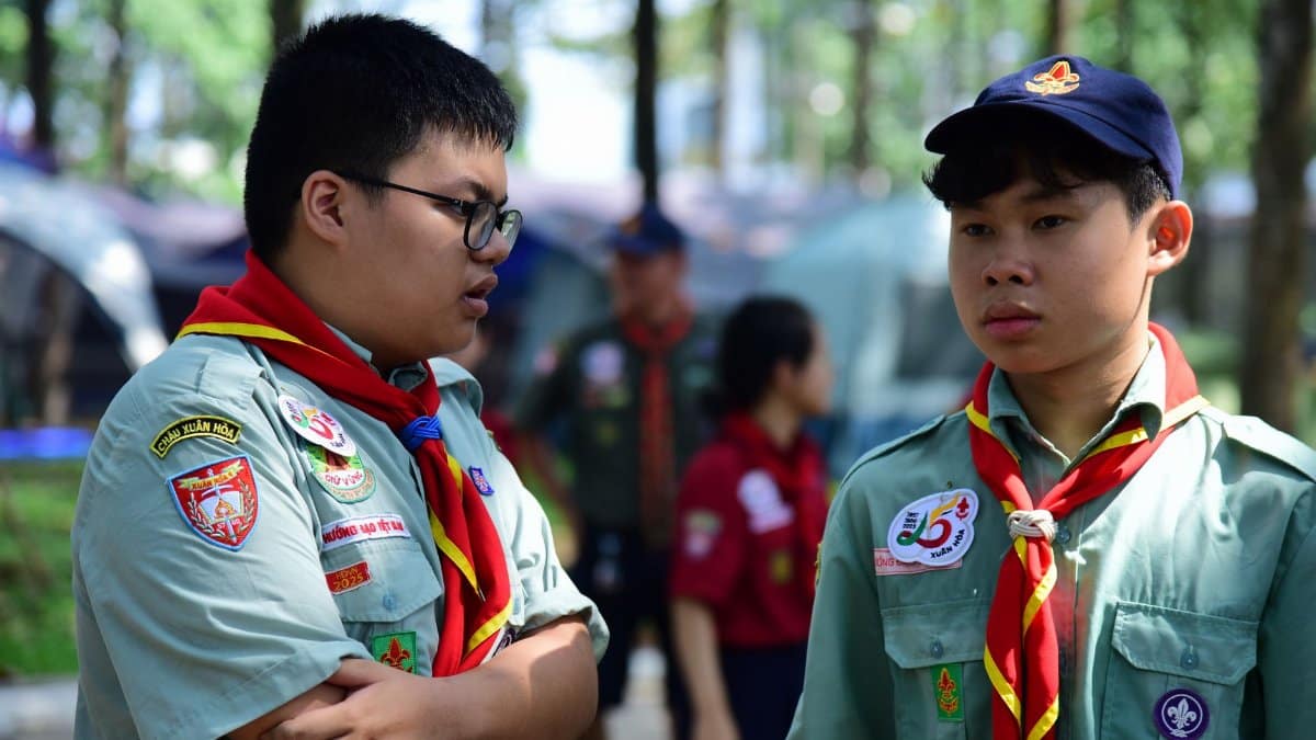 Two young scouts in uniform having a conversation at an outdoor camp, surrounded by tents and trees.