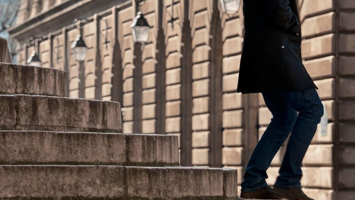 A person in jeans and trench coat ascending city steps beside historic buildings.