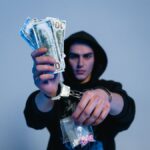 Handcuffed man in a hoodie holding cash and drugs, depicting crime and addiction.