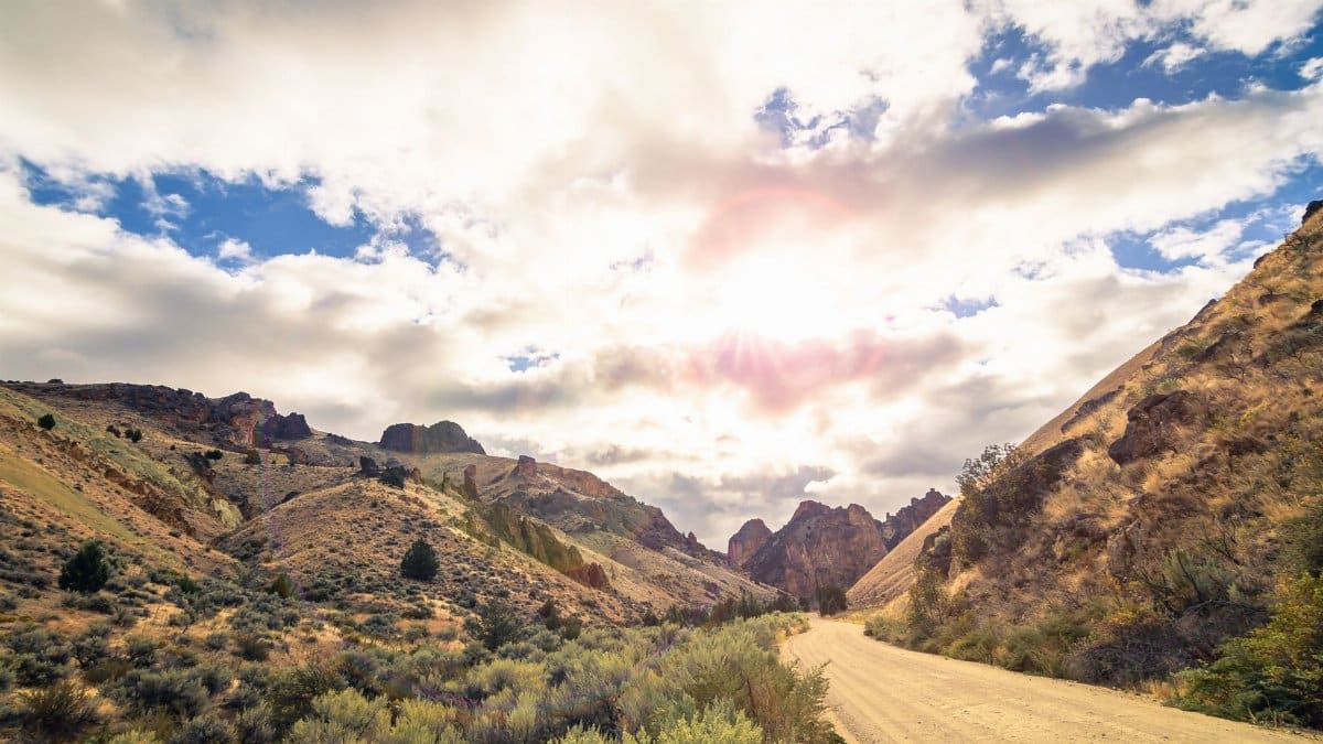 Picturesque scenery of empty sandy road running through rocky hills covered with dry grass and bushes under cloudy sky in daytime